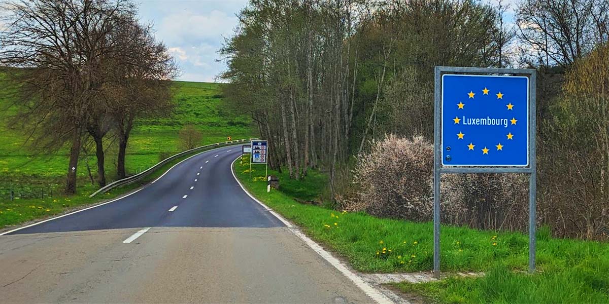 A two-lane highway curves through a green meadow with leafless trees on both sides. A road sign that reads "Luxembourg" inside the European Union flag is on the right.