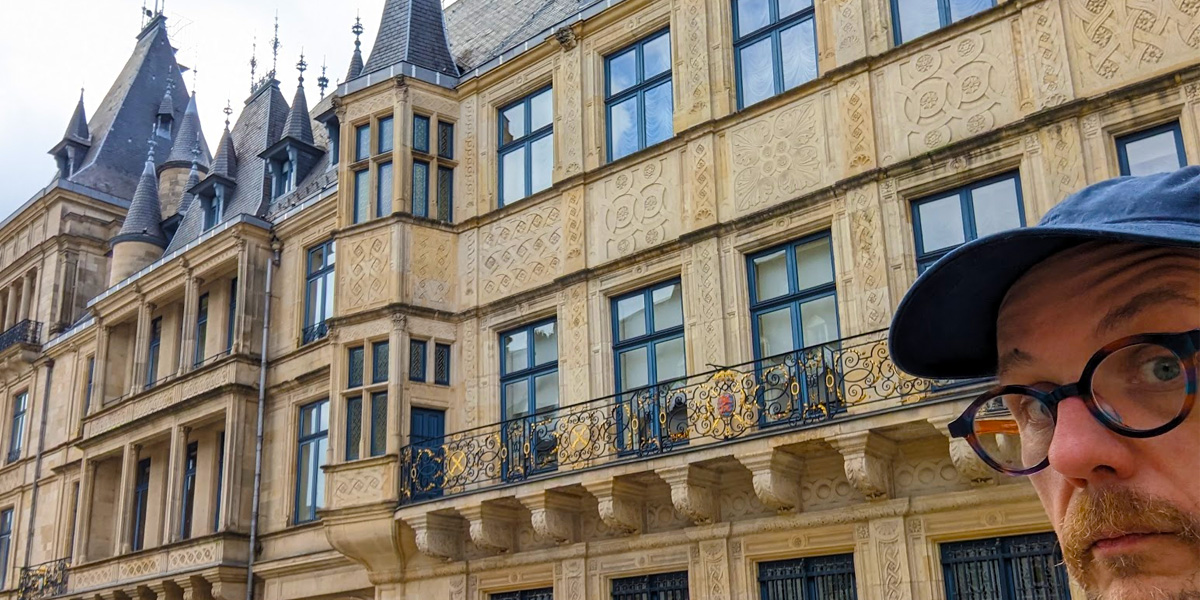 A white man wearing glasses and a baseball cap looks toward the camera from the lower right corner. The Grand Ducal Palace in Luxembourg City is behind him.